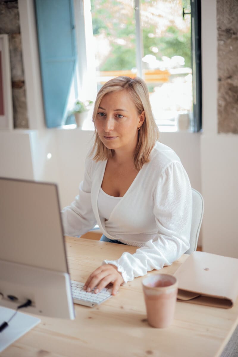 Frau am Schreibtisch mit Laptop und Kaffeebecher, helles Büro mit Fenster