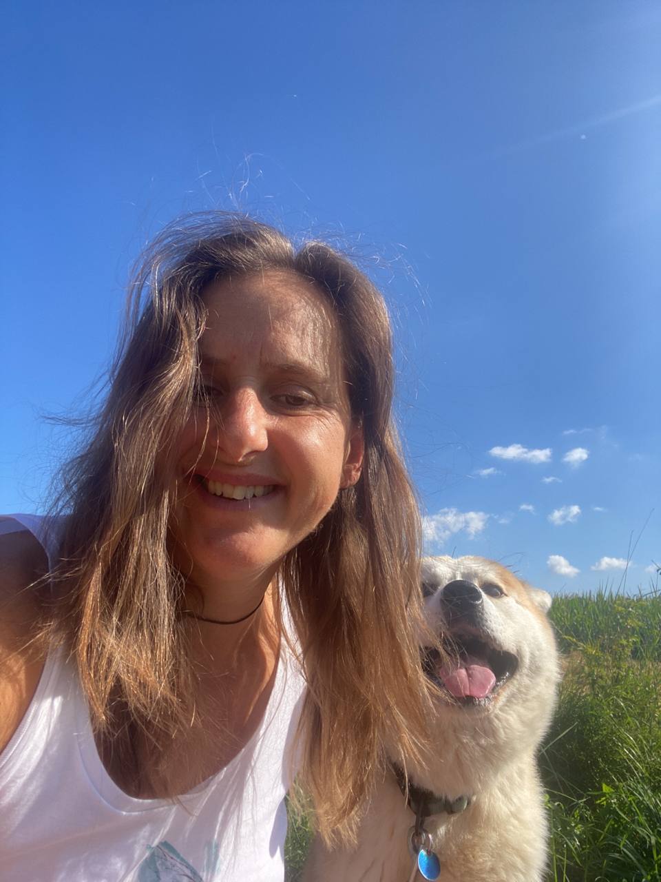 Frau mit braunen Haaren und Hund auf grüner Wiese unter blauem Himmel, lächelnd