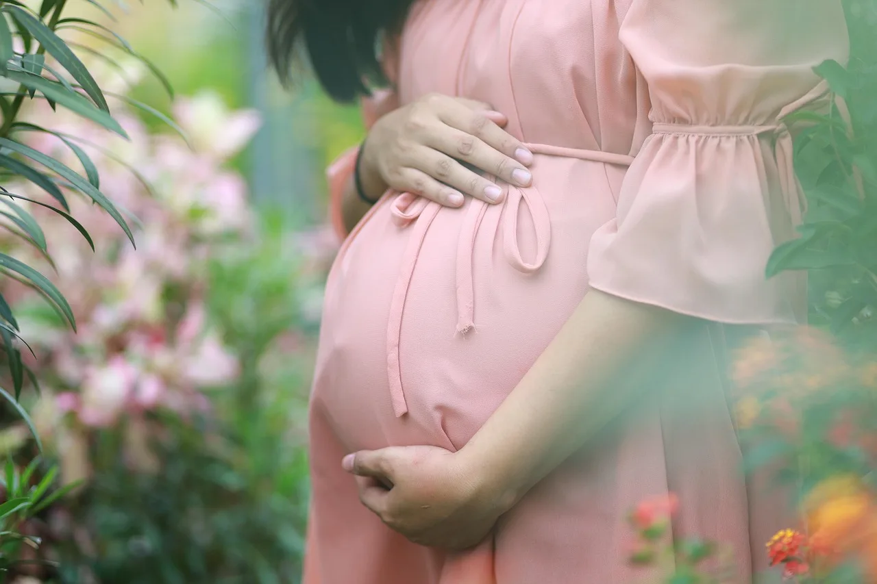 Schwangere Frau in rosa Kleid hält Bauch in Garten mit Blumen