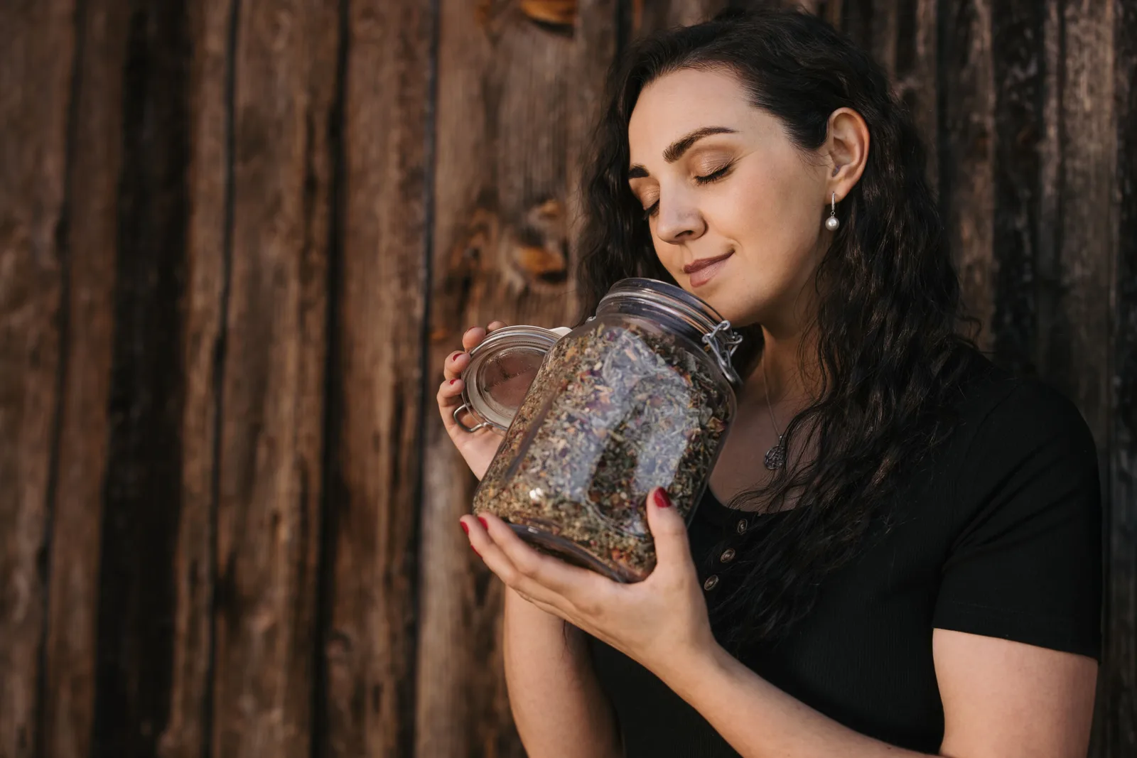 Selbstlernkurs Aromapraktiker*in von Manuela Hafner-Weber 3 Frau hält Glasgefäß mit Kräutermischung vor Holzwand, Fachwissen Aromatherapie