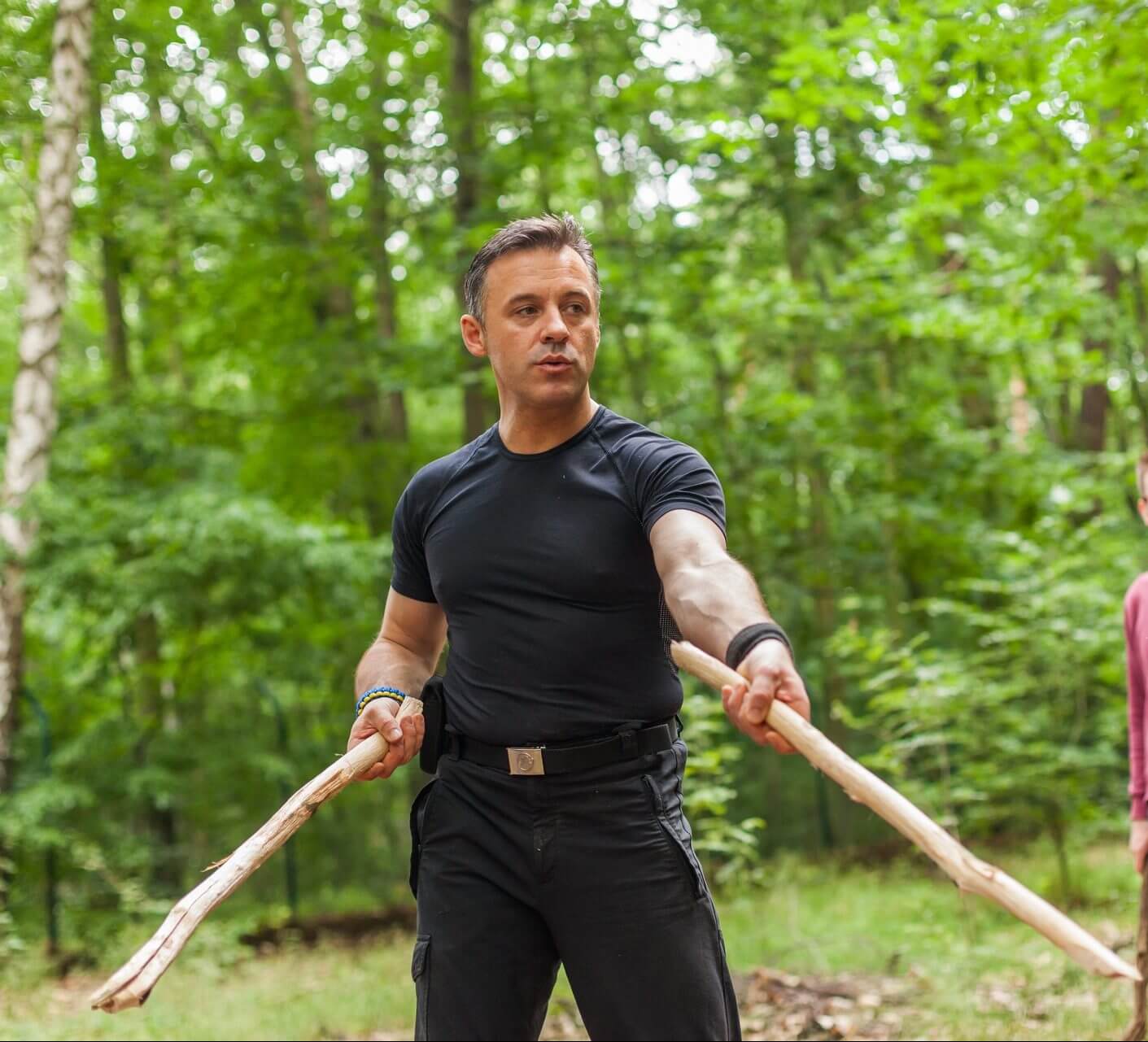 Selbstschutz & Verteidigung – Kurs zur Notwehr von Daniel Schäfer 8 Mann in schwarzem Trainingsanzug mit Holzstäben im grünen Wald