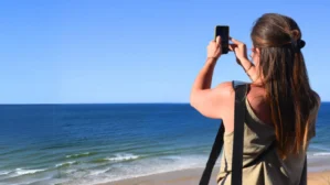 Frau fotografiert mit Smartphone am Strand vor blauem Himmel und Meer