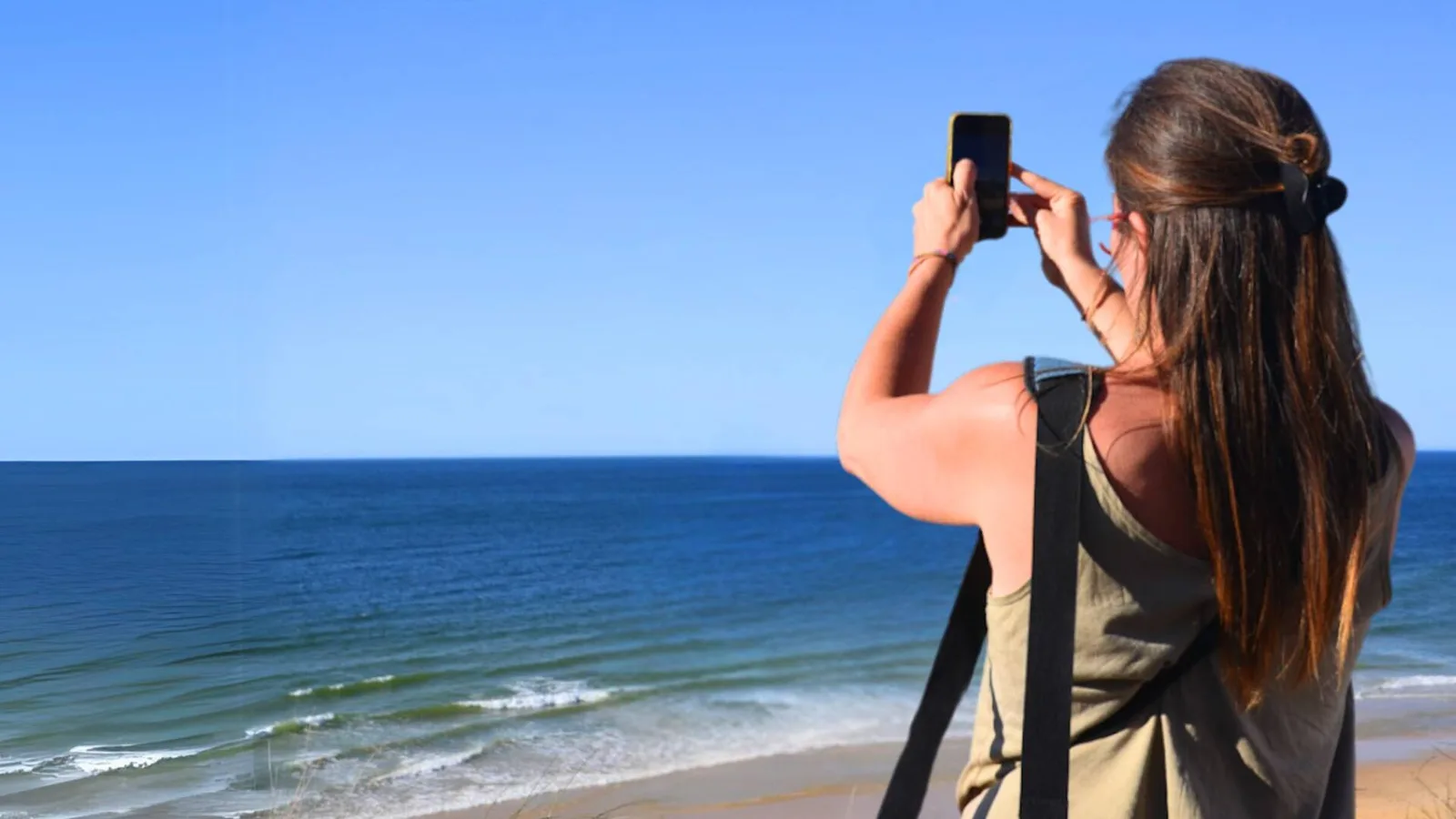 Frau fotografiert mit Smartphone am Strand vor blauem Himmel und Meer