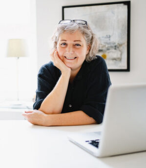 Porträt Constanze Straub am Schreibtisch mit Laptop, lächelnd, Hand am Kinn, helles Büro