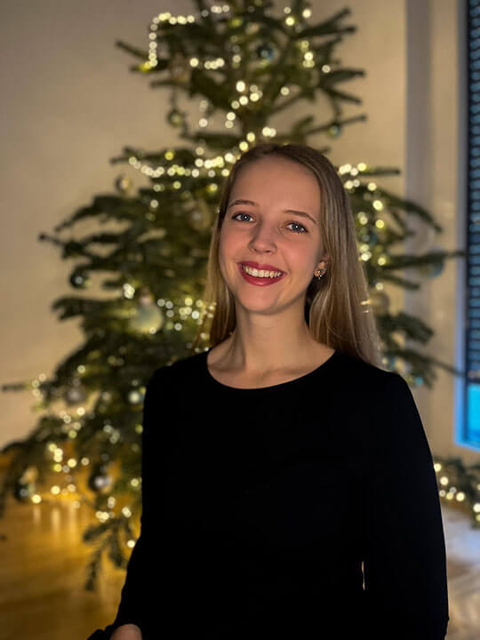 Jaqueline lächelnd vor beleuchteter Weihnachtstanne, schwarzes Oberteil