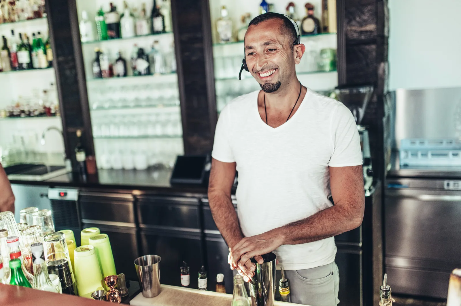 Barkeeper in weißem T-Shirt mit Kopfset an professioneller Cocktailbar, gut ausgestattete Flaschenregale im Hintergrund