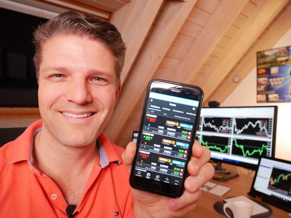 Frédéric Ebner in rotem Hemd zeigt Smartphone mit Tradingcharts, im Hintergrund Monitore mit Börsendiagrammen