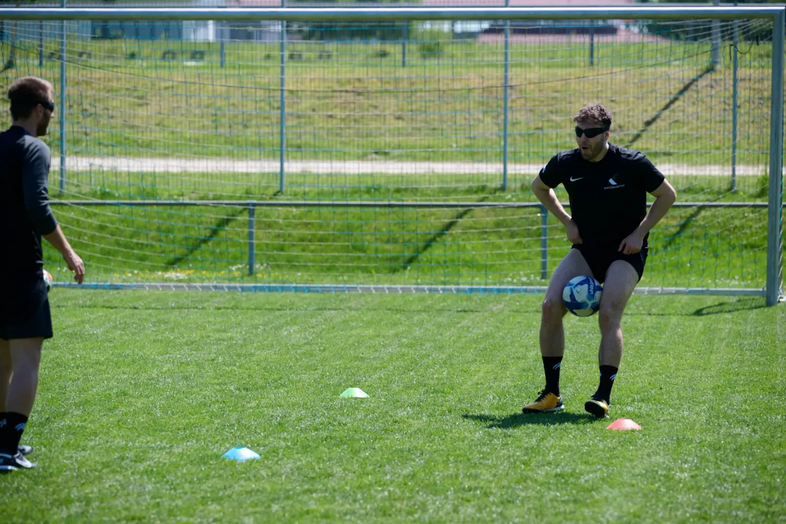Fußballtraining auf Rasenplatz mit Torwart in schwarzem Nike-Trikot, der den Ball kontrolliert, während ein Trainer zuschaut