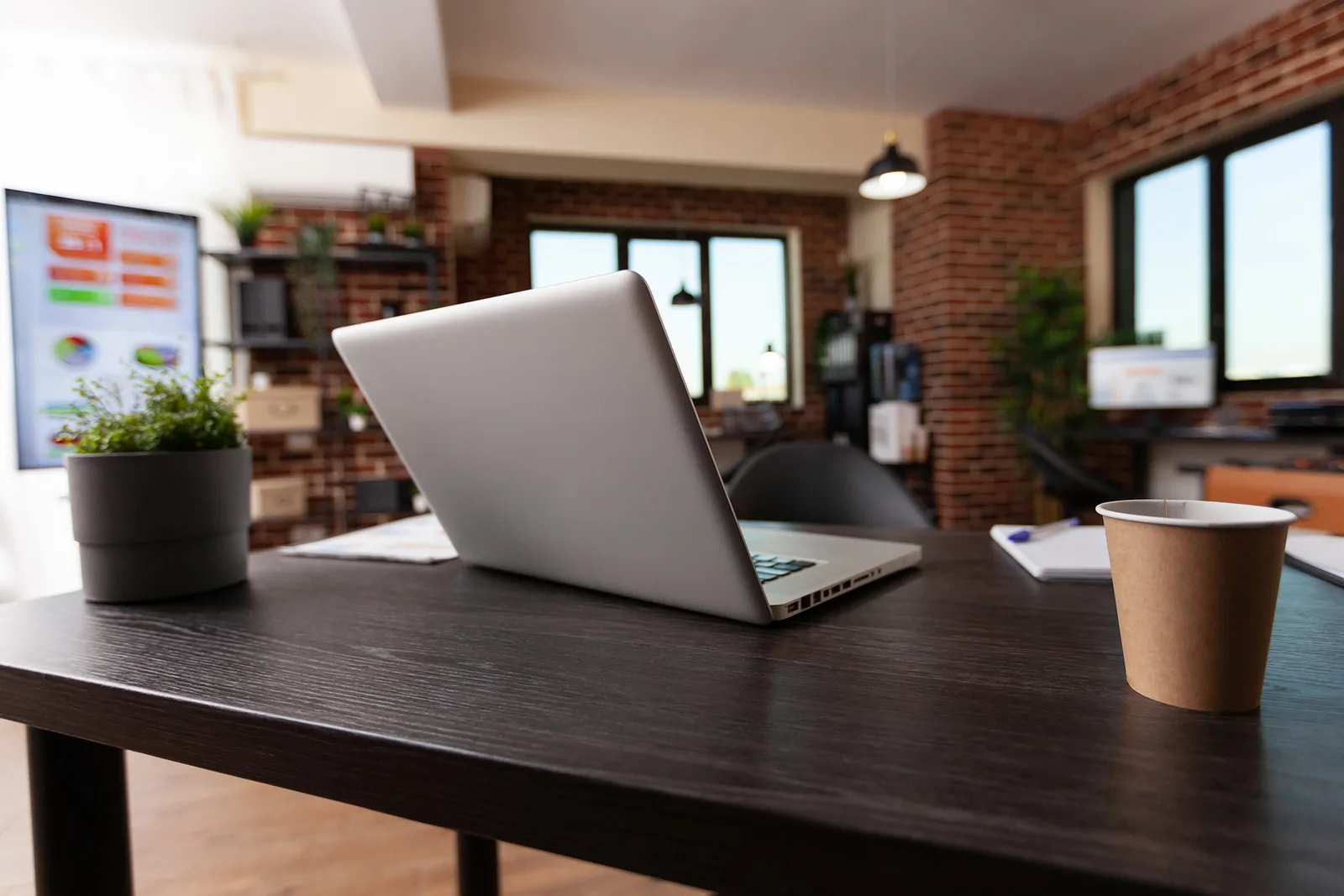 Modernes Büro mit Laptop auf dunklem Tisch, Pflanze, Kaffebecher, Ziegelmauer und Fenster im Hintergrund