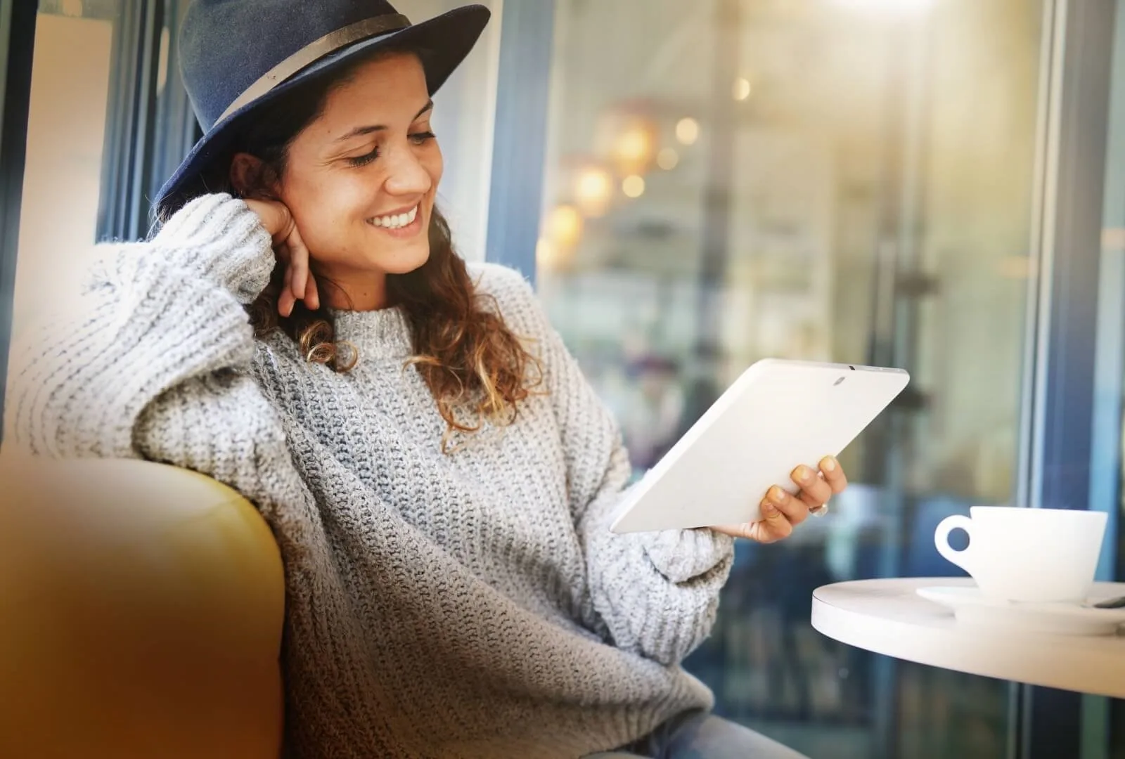 Lachende Frau mit Hut liest auf Tablet am Fenster mit Kaffee