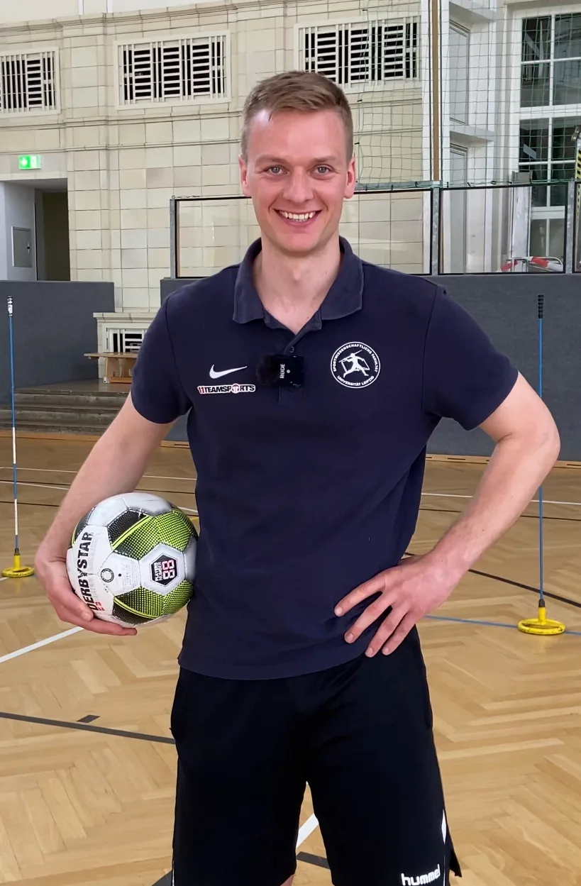 Erich Reich, Trainer in dunklem Poloshirt, hält Fußball in Sporthalle, Lächeln