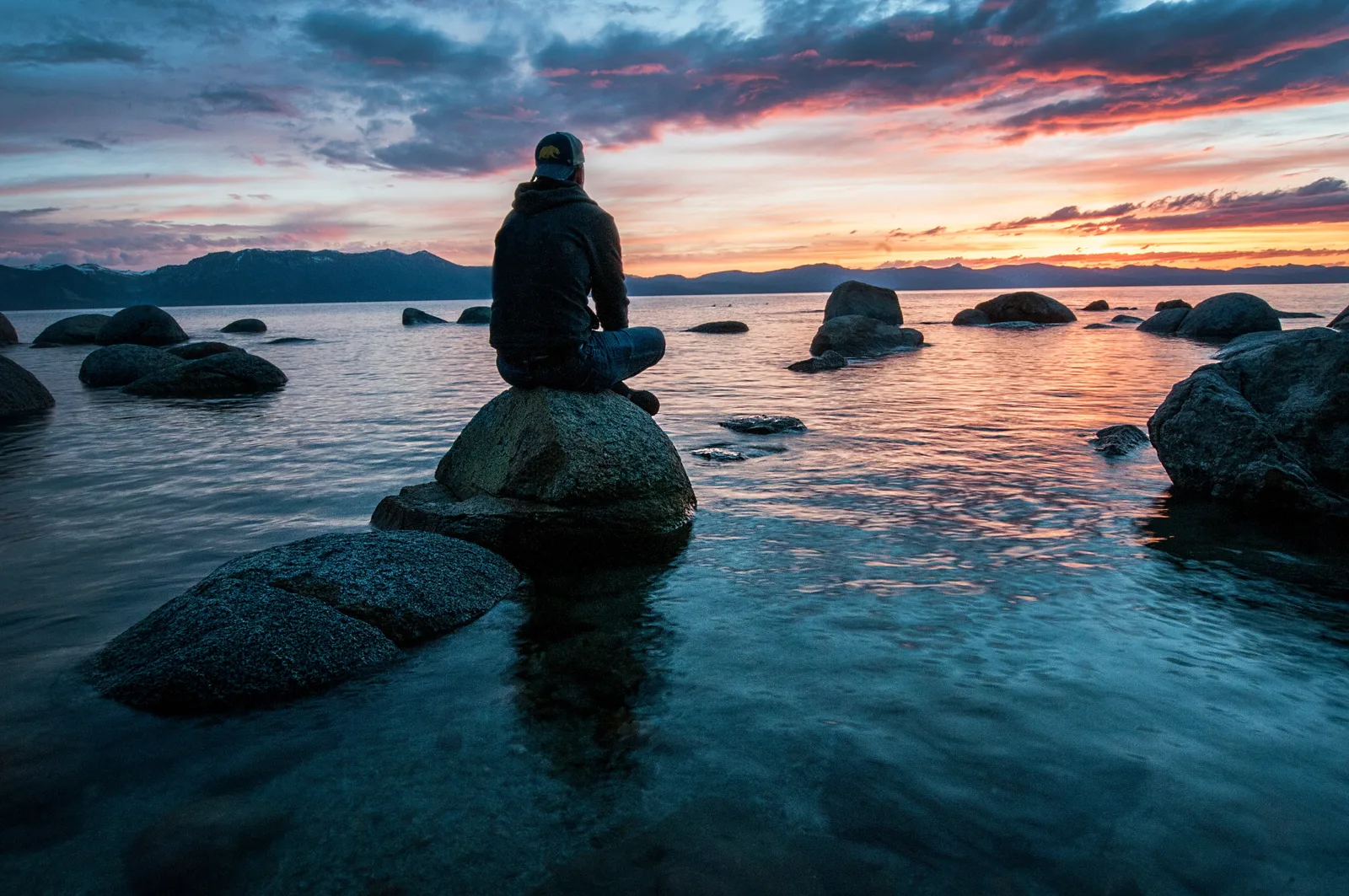 Stark im Kopf Kartenset – 50 Übungskarten für mentales Training 4 Person sitzt auf Stein im Wasser, blickt auf See mit Bergen und dramatischem Himmel bei Sonnenuntergang