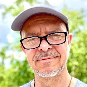 Porträtfoto von Torsten Zumpe mit Brille und grauer Mütze vor grünem Hintergrund