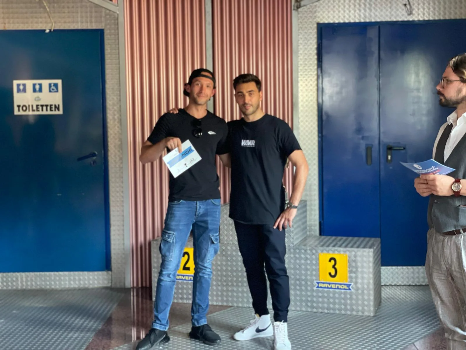 Zwei Rennfahrer auf Siegertreppchen mit Zertifikaten und Person in Businesskleidung