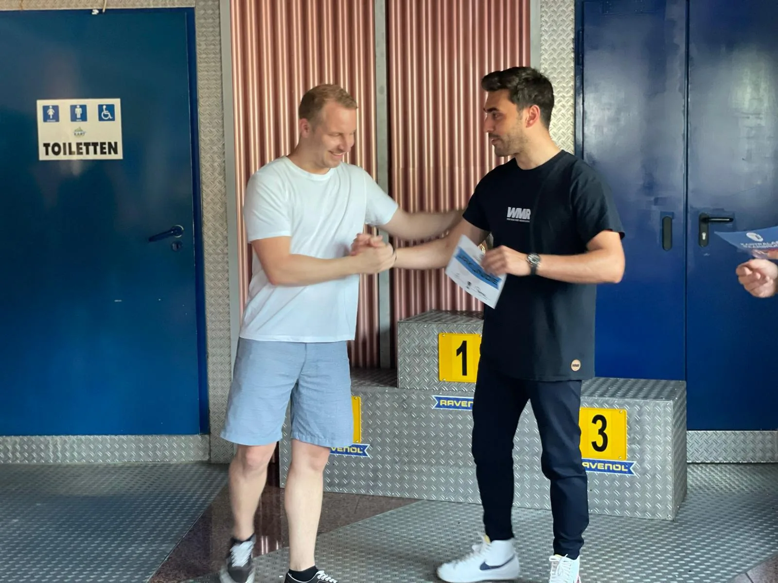 Zwei Männer bei Übergabe eines Zertifikats vor Podium mit gelben Nummernblöcken