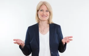 Insa Künkel, blonde Frau in dunklem Blazer mit weißer Bluse, offene Handflächen, Lächeln