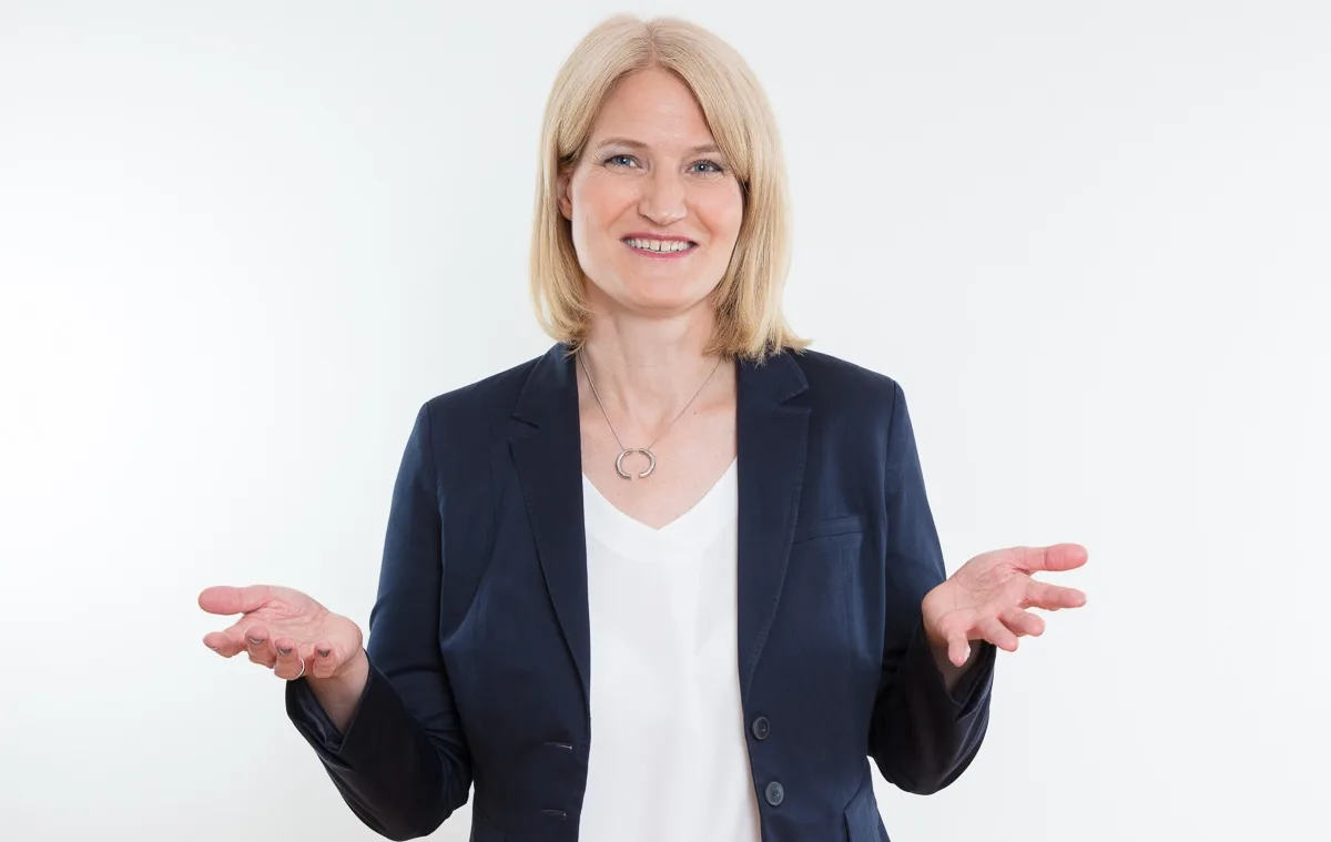 Story klar – Erzählcoaching für Experten von Insa Künkel 1 Insa Künkel, blonde Frau in dunklem Blazer mit weißer Bluse, offene Handflächen, Lächeln
