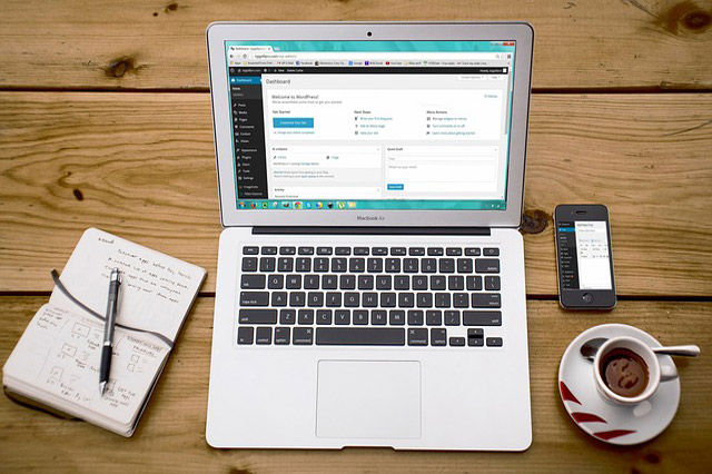 Laptop mit WordPress-Dashboard auf Holztisch, Notizbuch, Stift, Smartphone und Kaffeetasse