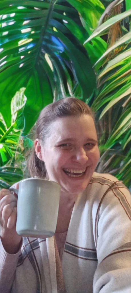 Rabea Baierl von Systemgedacht lacht mit Kaffeetasse vor grüner Pflanze