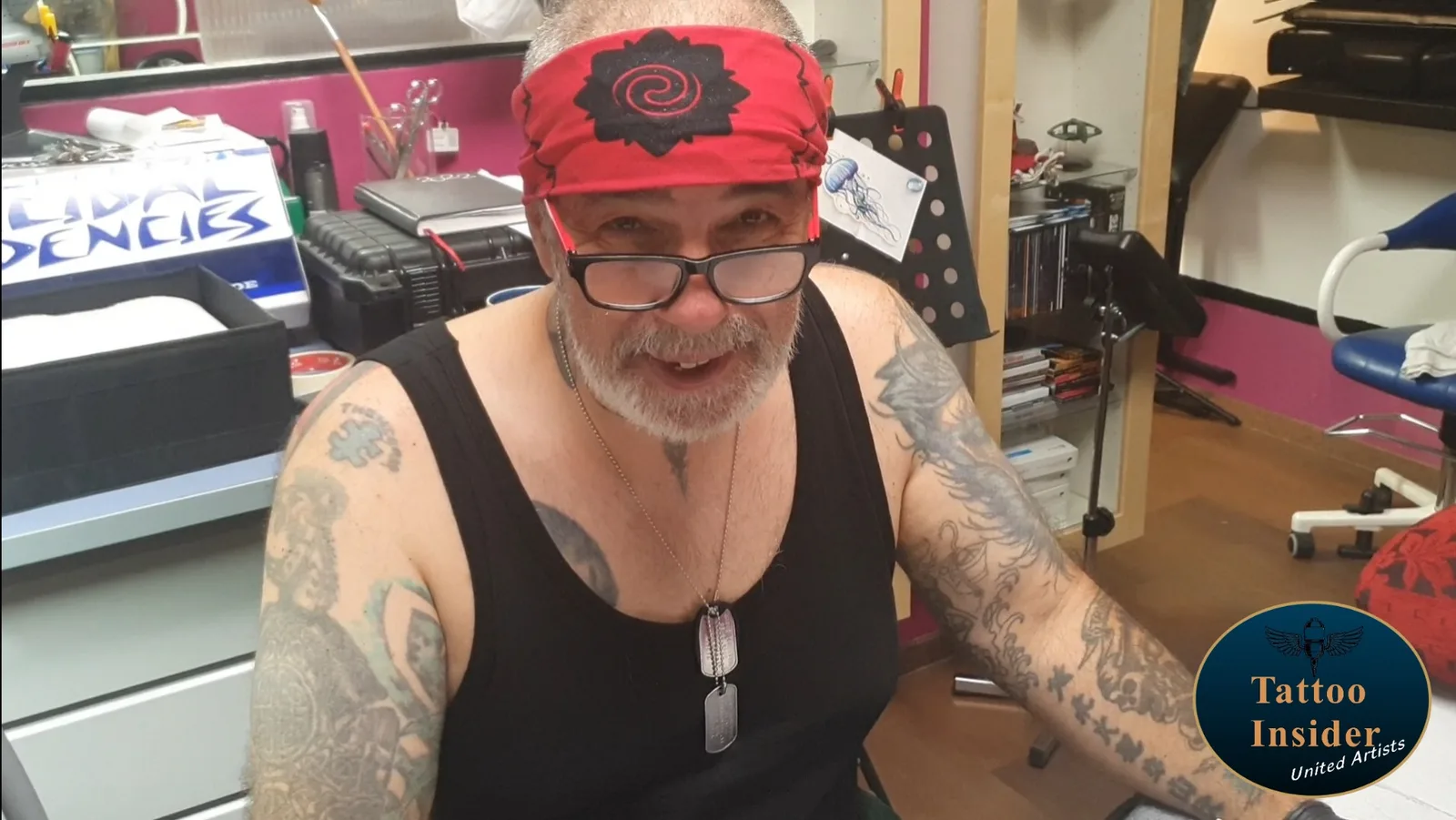 Tattoo-Artist mit roter Mütze im Studio, trägt schwarzes Shirt und Kettenhalskette, lächelt in die Kamera