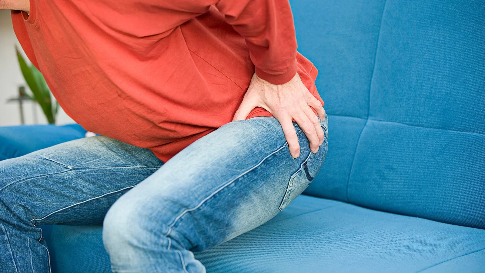 Person in rotem Oberteil sitzt auf blauem Sofa und fasst sich an das Hüftgelenk