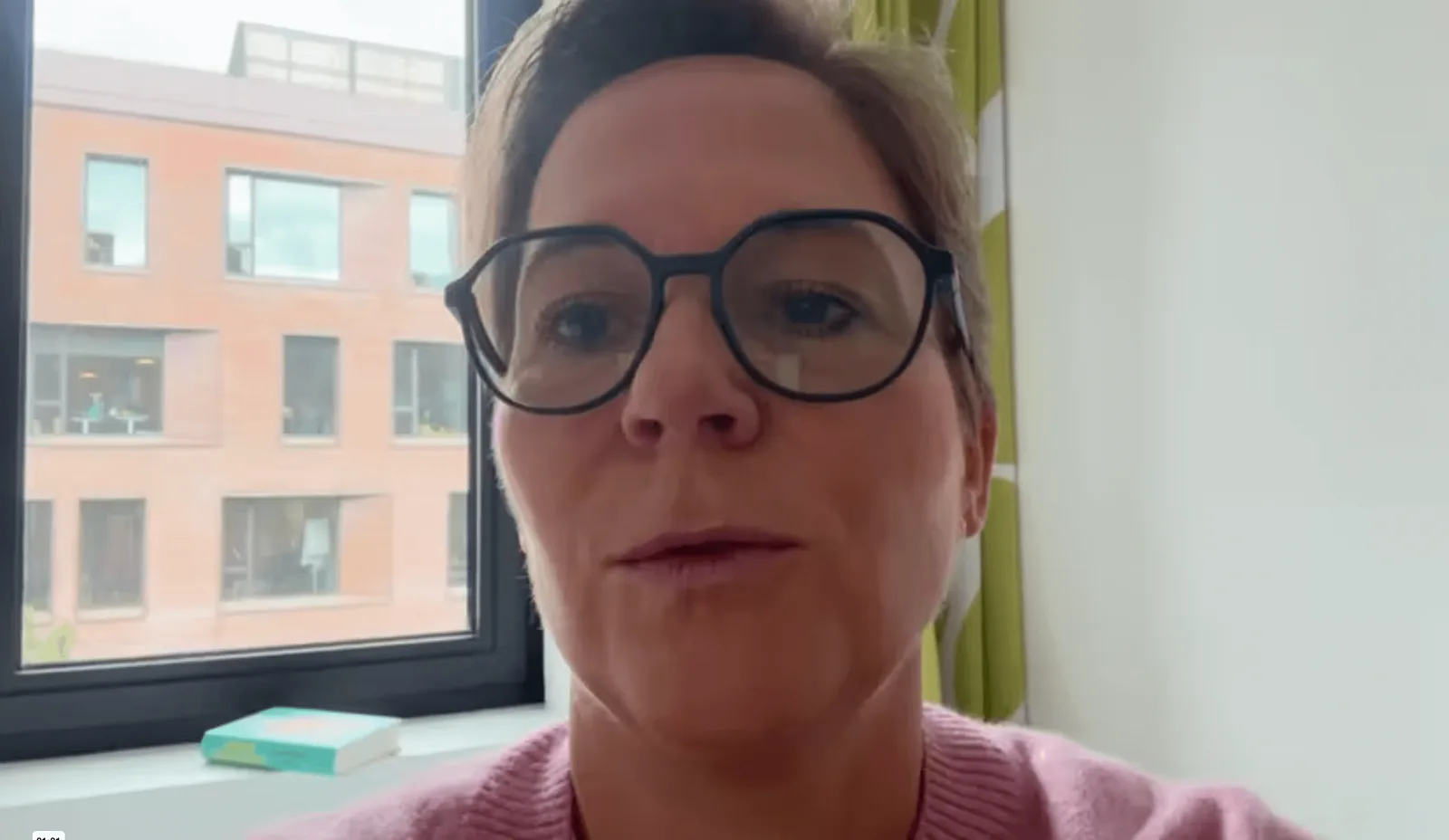 Videostandbild Katja Hinterleitner Selfie vor Fenster und Gebäude, trägt Brille