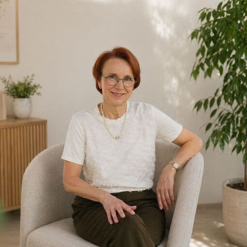 Gina Heller sitzt in grauem Sessel, weißes Shirt, helles modernes Wohnzimmer mit Pflanzen