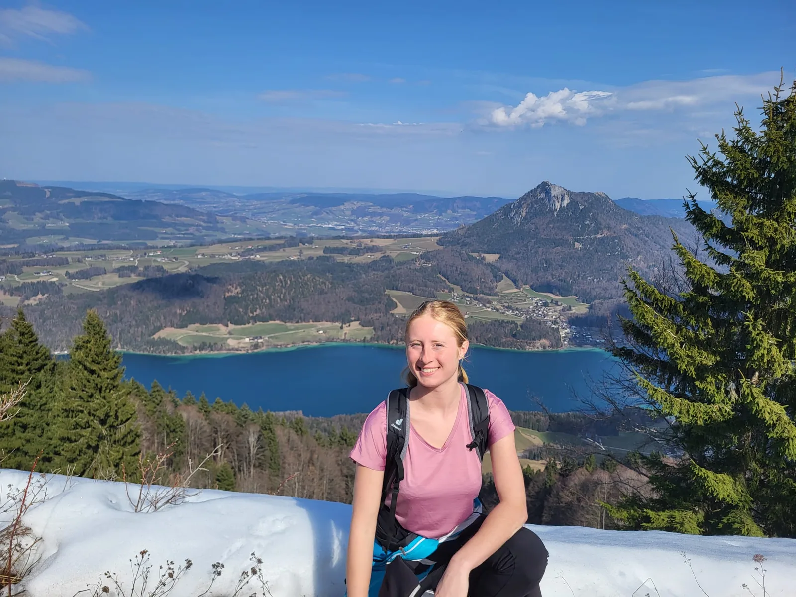 Junge Frau in rosa Shirt mit Rucksack sitzt vor Bergsee und grünem Tal