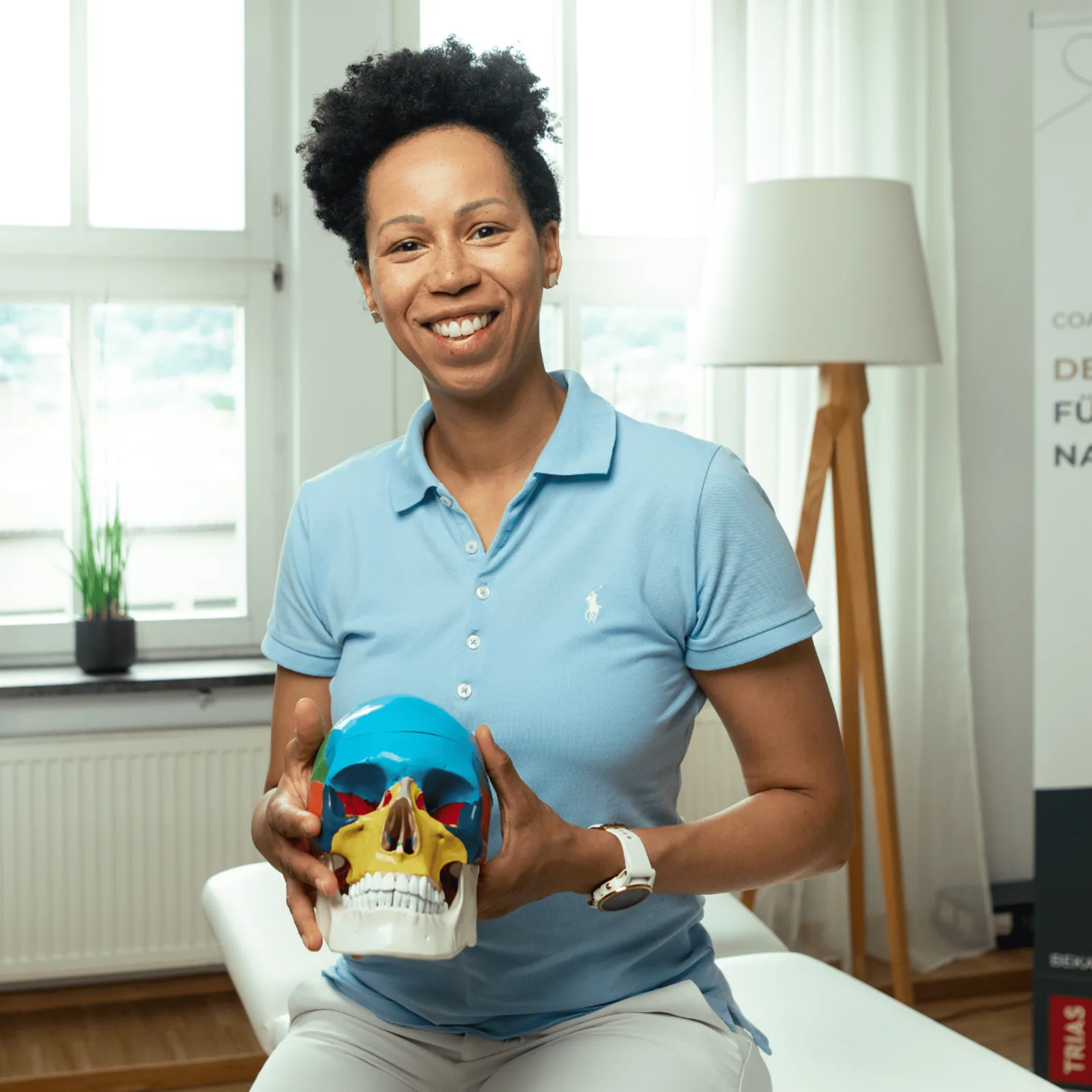Frau mit schwarzen Haaren in hellblauem Poloshirt hält farbiges Anatomie-Schädelmodell, modern eingerichteter Raum