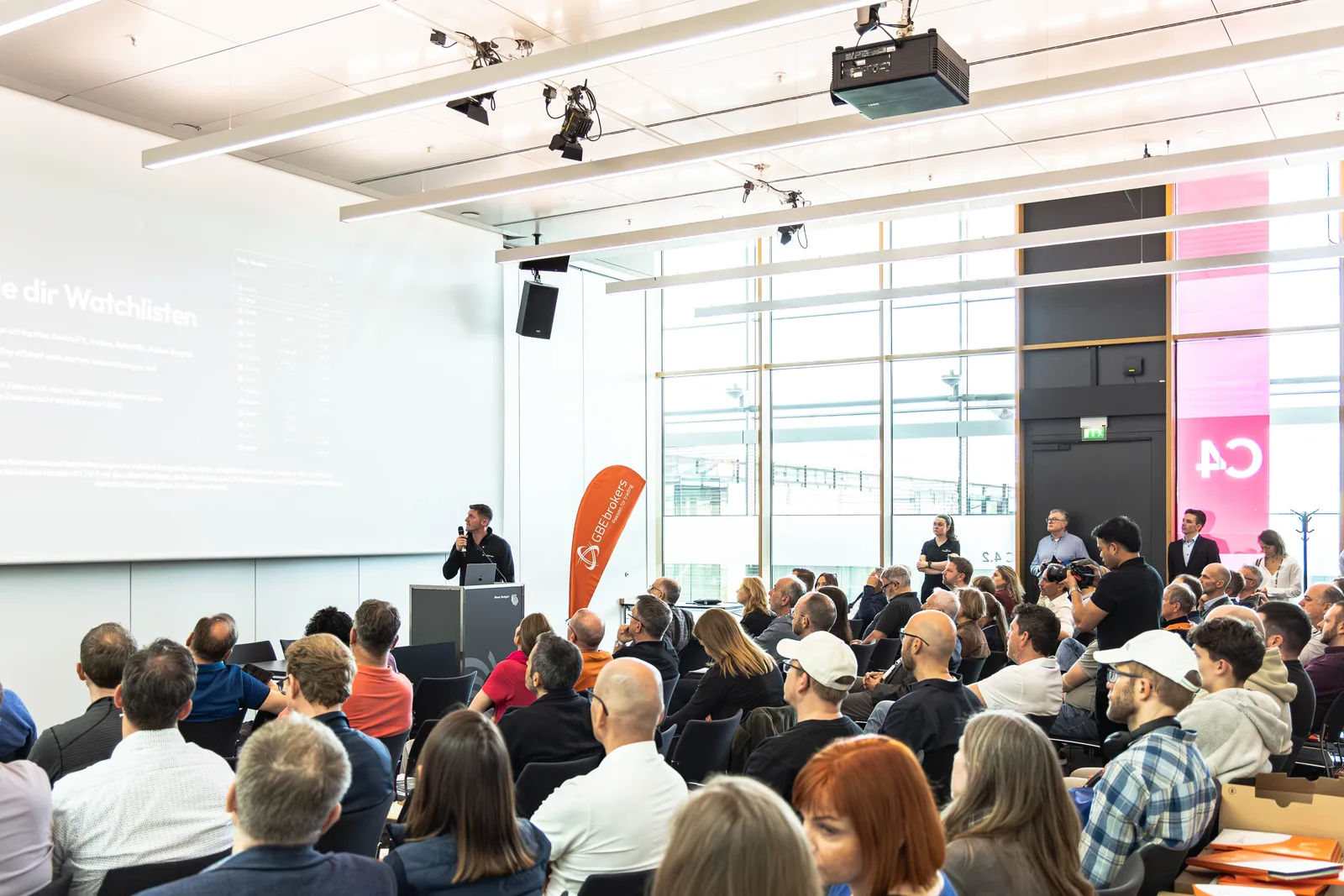 Konferenzraum mit Publikum bei Präsentation, Referent am Podium spricht zu Zuhörern, großes Fenster mit Stadtblick
