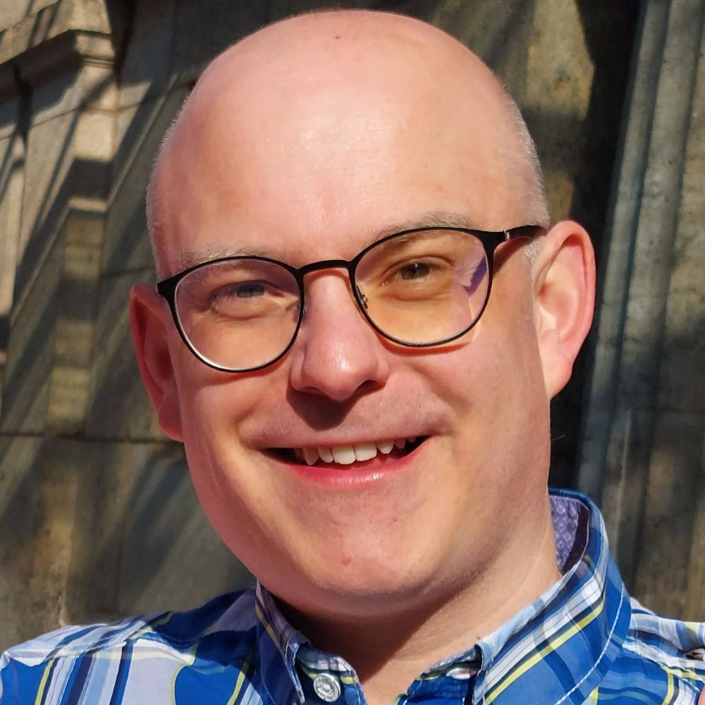 Porträt eines lächelnden Mannes mit Brille und kariertem Hemd vor unscharfem Hintergrund