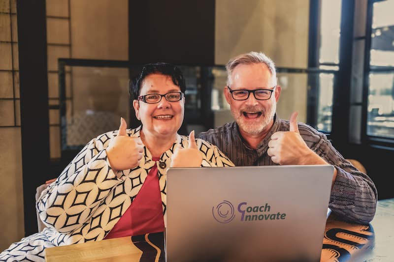 Zwei Personen vor Laptop mit Coach Innovate Logo machen Daumen-hoch-Geste