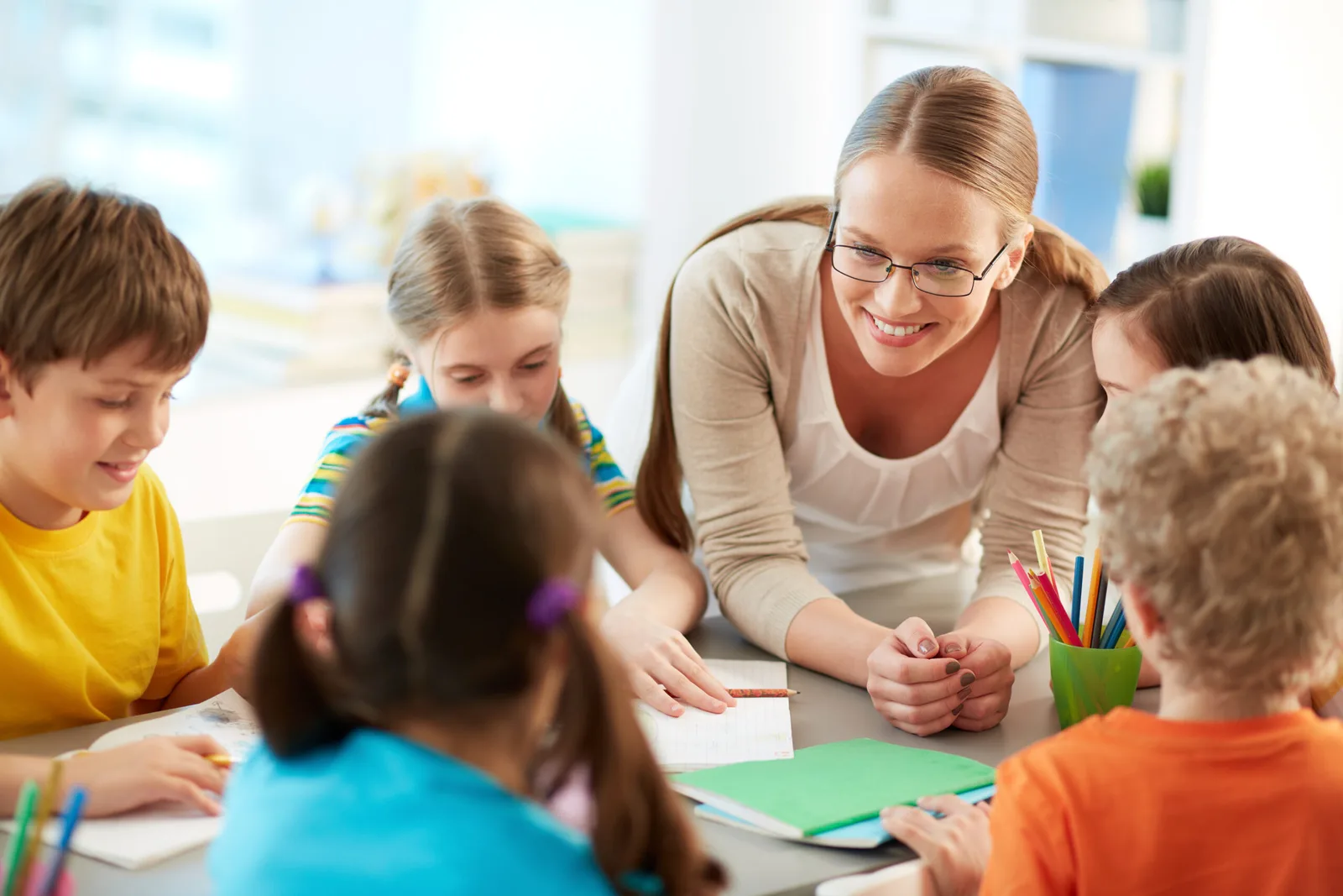 Lehrerin mit Brille sitzt lächelnd mit vier Kindern am Tisch und hört ihnen zu