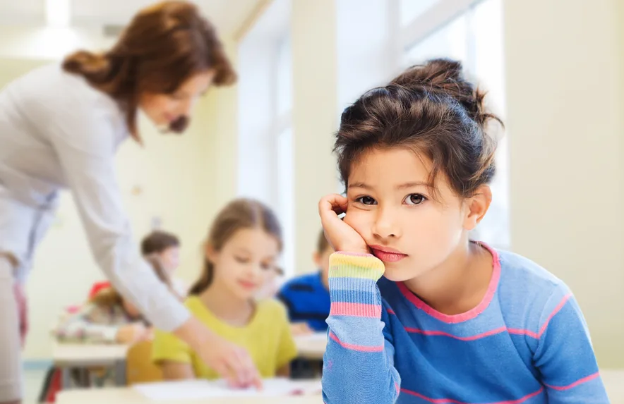 Mädchen in blauem Shirt sitzt nachdenklich mit aufgestütztem Arm, Lehrerin im Hintergrund mit anderen Kindern