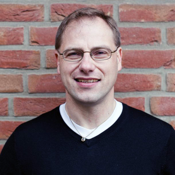 Porträt von Peter Schmidt, Mann mit Brille und schwarzem Pullover vor Backsteinmauer