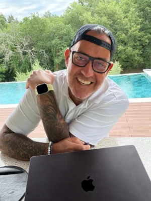 Ralf Schmitz am Pool mit Laptop, weißes Shirt, Brille, goldene Smartwatch, lächelnd
