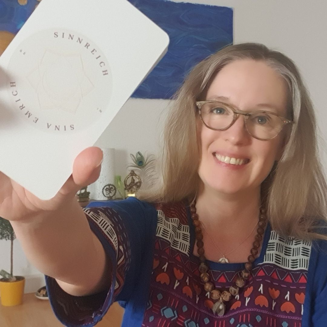 Visionskarten Atelier – Videokurs zur Zielklarheit von Sina Seybold 3 Frau mit Brille und Zöpfen hält lächelnd Buch 'SinnReich' vor blauem Hintergrund, Dekoration mit kleinen Pflanzen