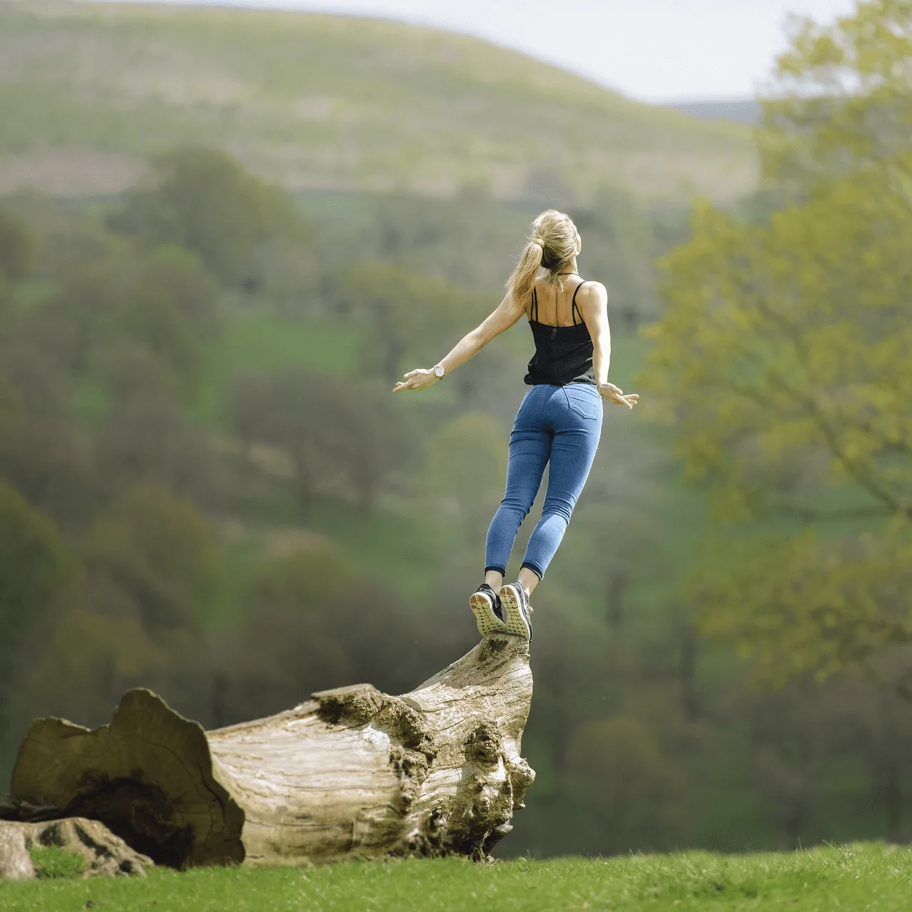 Frau mit blondem Haar springt von Baumstamm in grüner Landschaft mit Wald im Hintergrund
