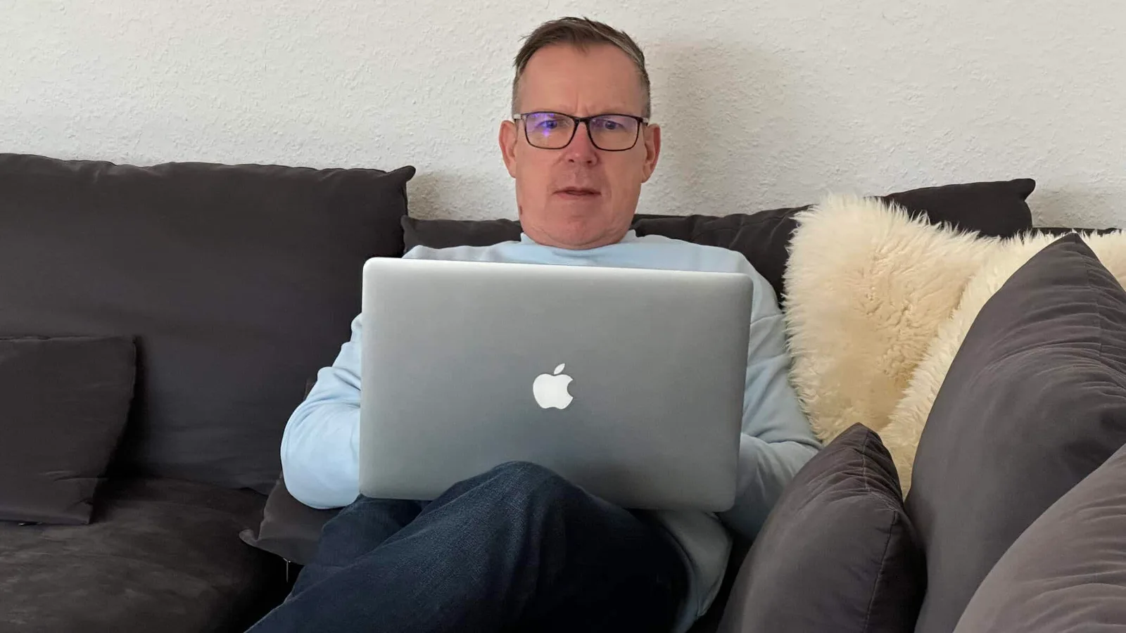 Voice to Mail – Web-App zur Sprachdokumentation von Michael Laschitza 1 Michael Laschitza sitzt auf schwarzem Sofa mit MacBook, trägt Brille und hellblaues Shirt