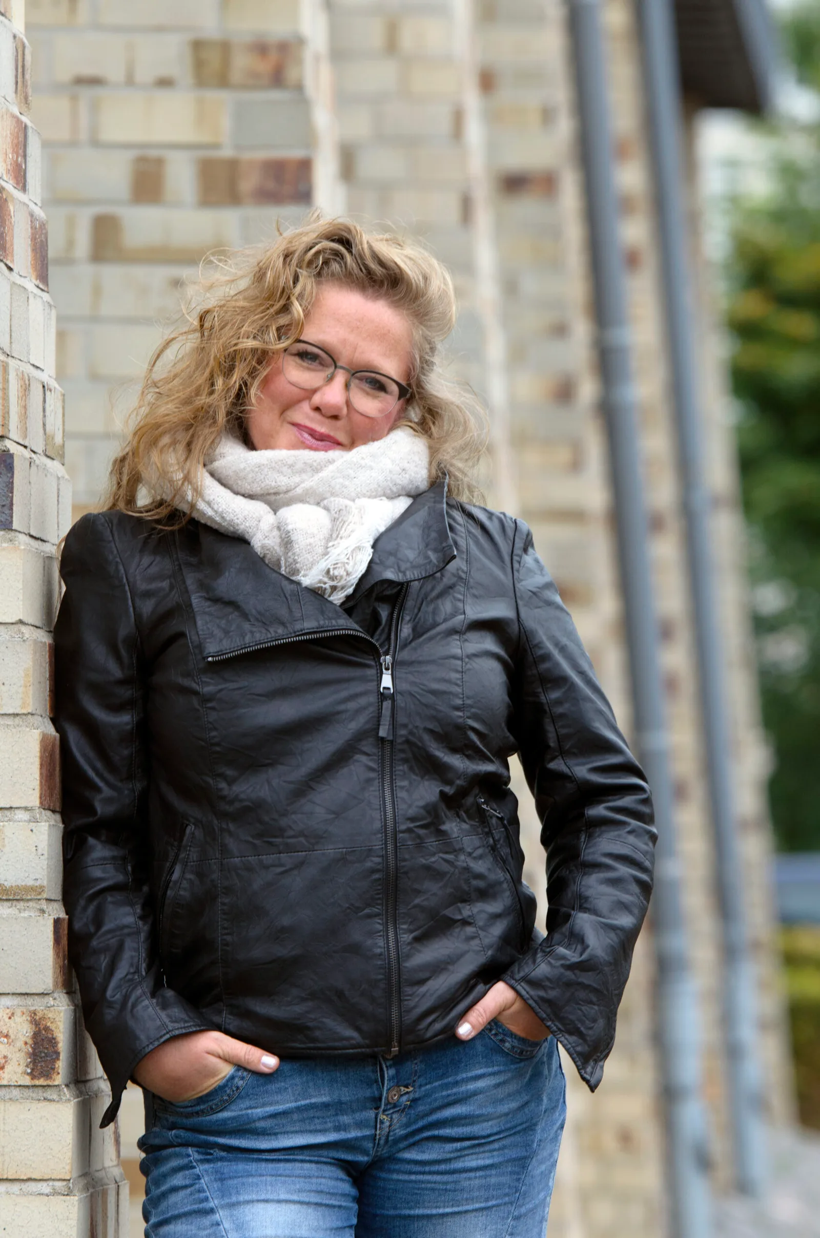 Tanja Rossis an Backsteinmauer lehnend, schwarze Lederjacke, weiß gewickelter Schal, urban outdoor