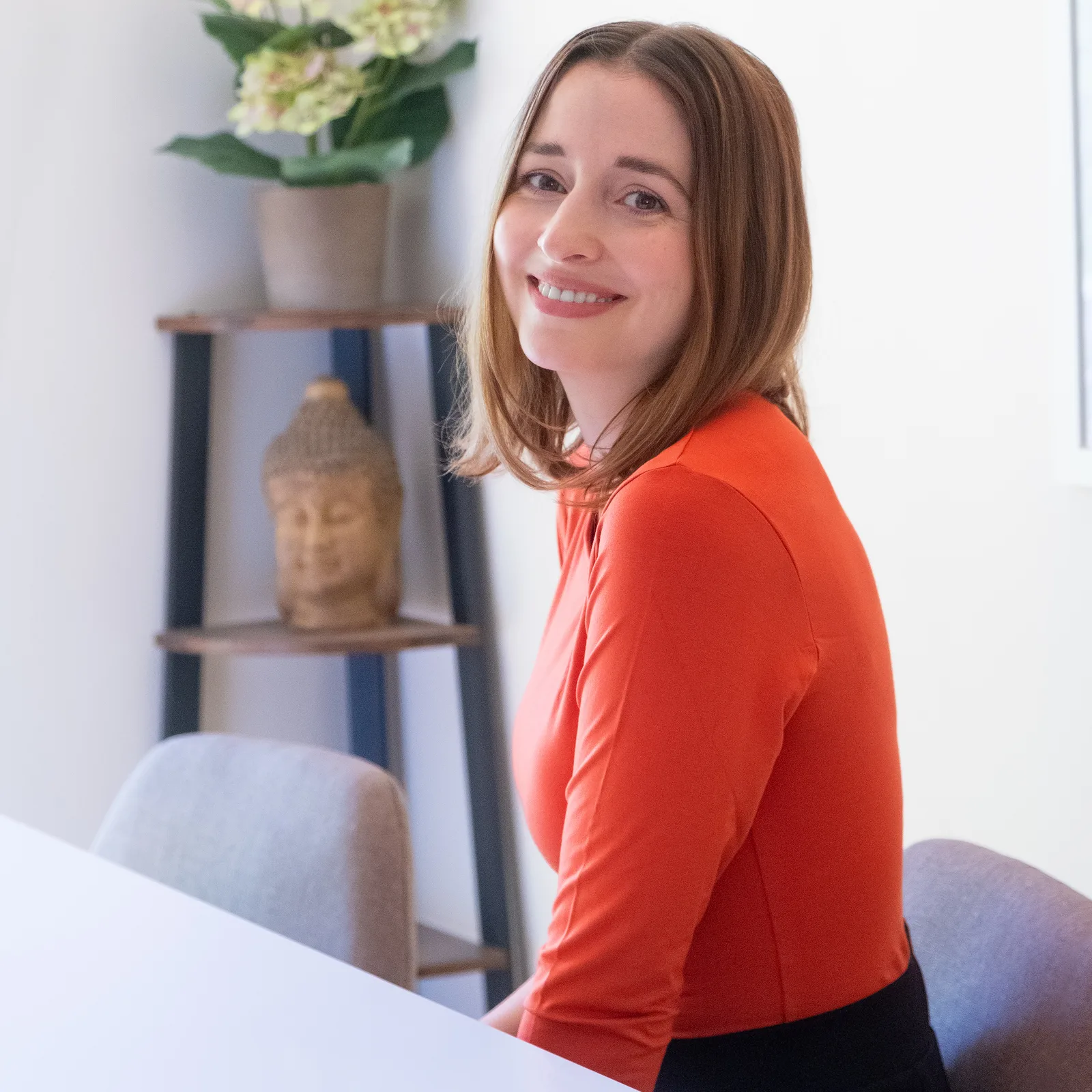 Portrait Dr. Katharina Tempel mit rötlichem Oberteil, sitzt vor Regal mit Buddha-Statue und Pflanze