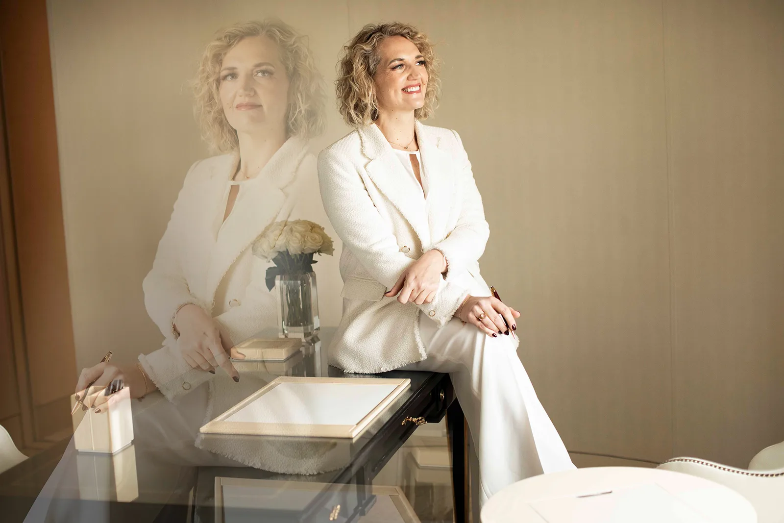 Christina Sternbauer in weißem Blazer sitzt am Schreibtisch mit Blumenvase und Notizbuch, im Hintergrund ihre Spiegelung