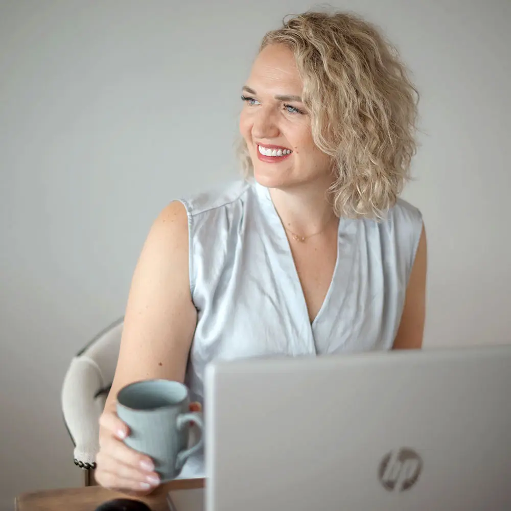 Christina Sternbauer lächelnd am Laptop mit Kaffeetasse, helles Bluse, seitliches Porträt