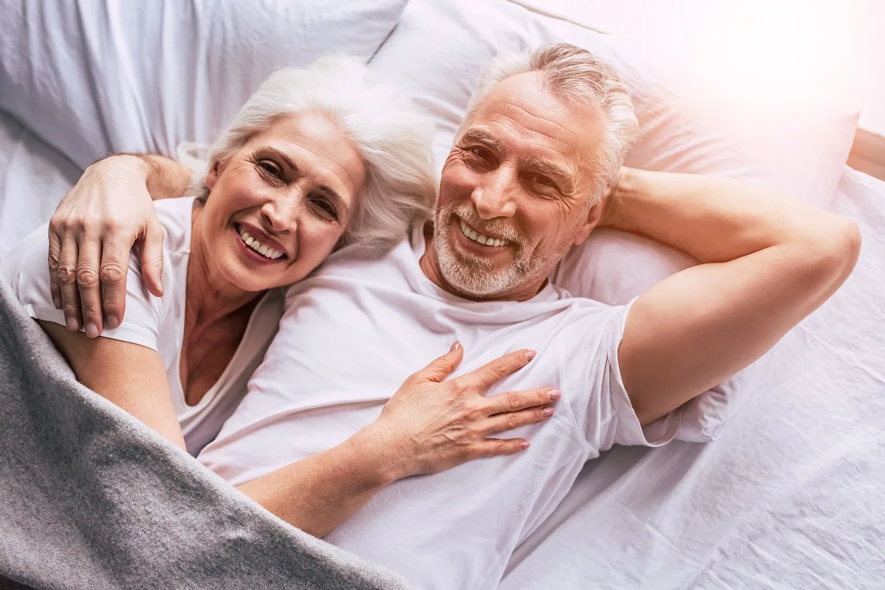 Glückliches älteres Paar lächelt im Bett und umarmt sich unter weißer Bettwäsche