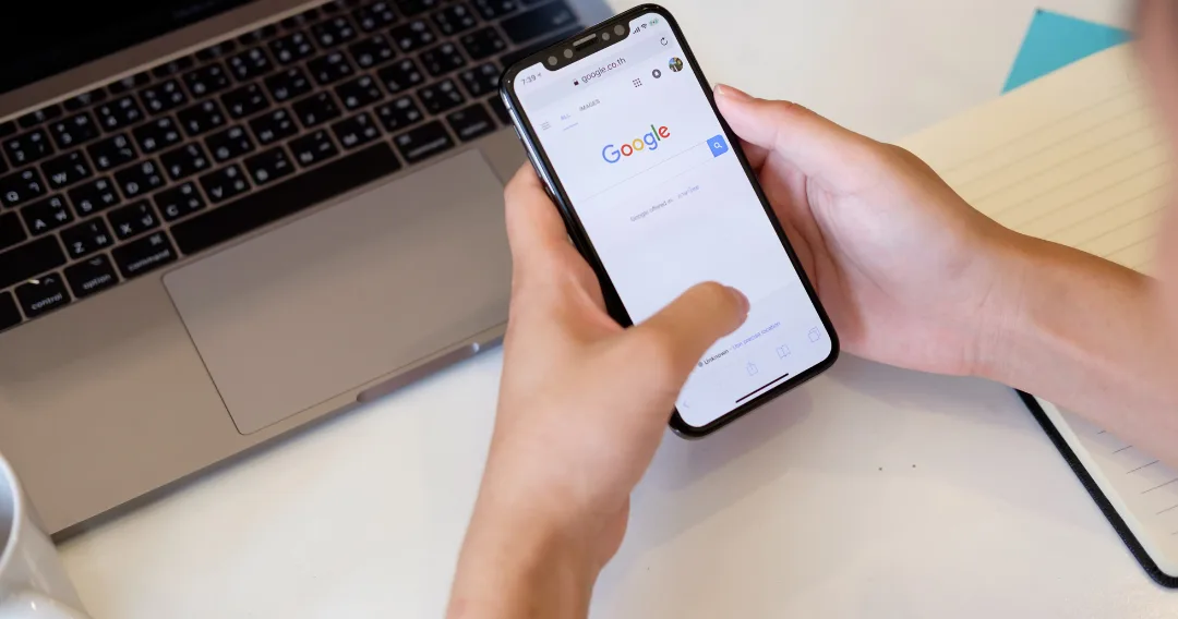 Hand hält Smartphone mit Google-Suchseite vor Laptop-Tastatur
