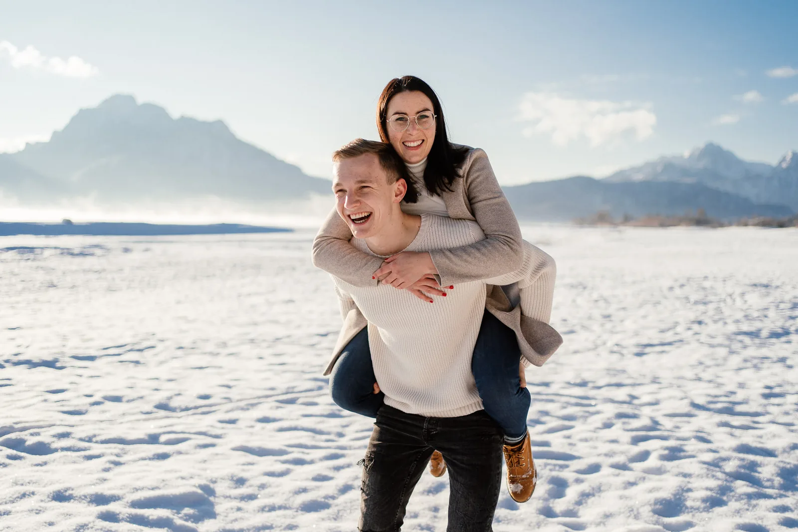 Junges Paar lacht am Strand in Bad Tölz vor Bergpanorama im Schnee bei Wintershooting