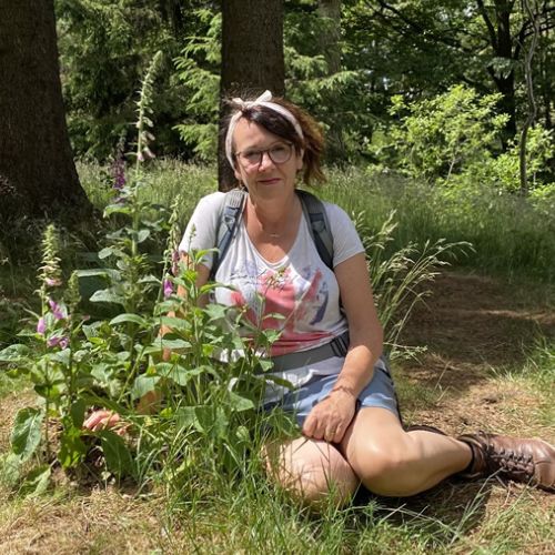 Simone Detto kniend im Wald neben Blumen, Porträtfoto mit Brille