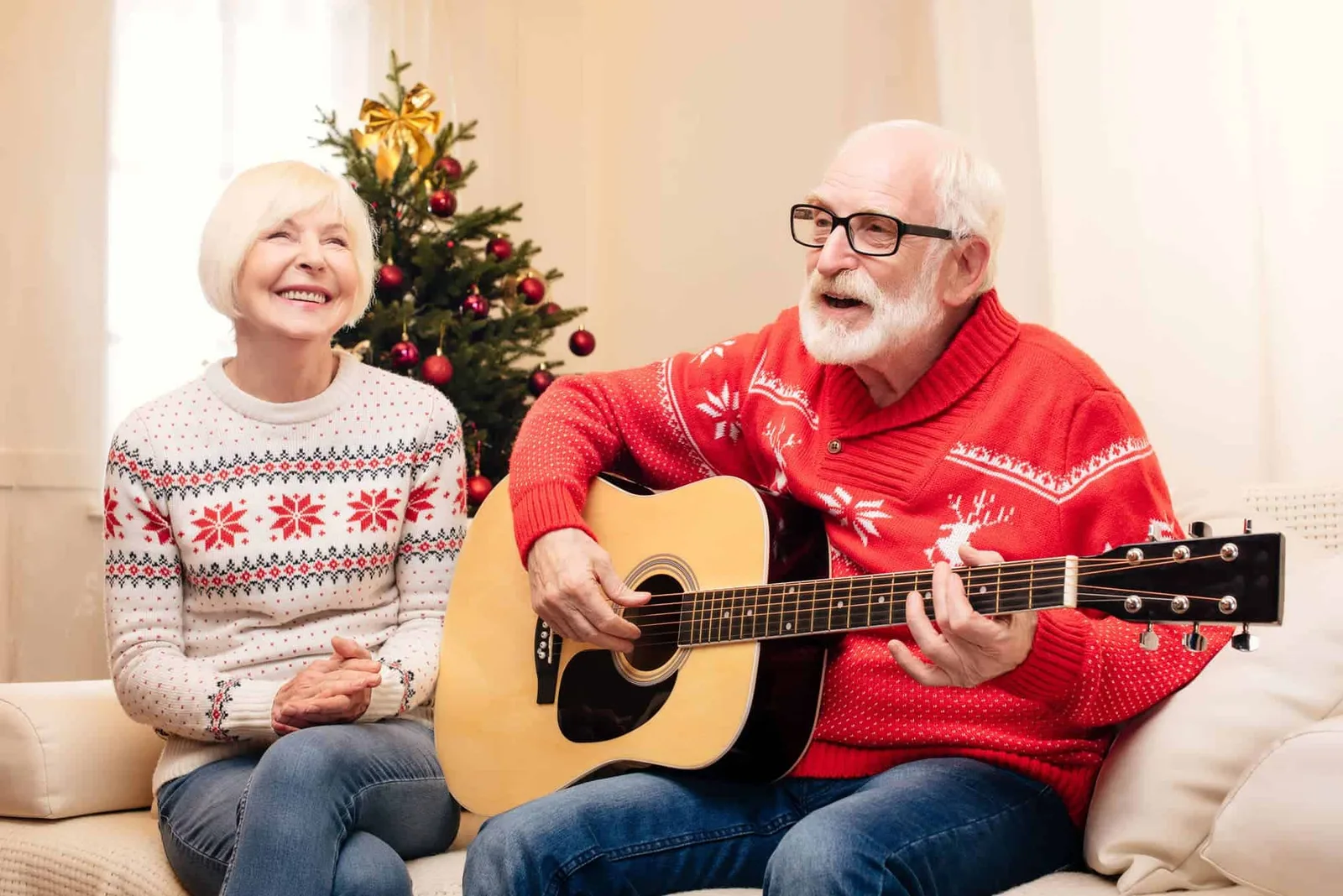 Älteres Paar in Weihnachtspullovern spielt Gitarre vor Weihnachtsbaum
