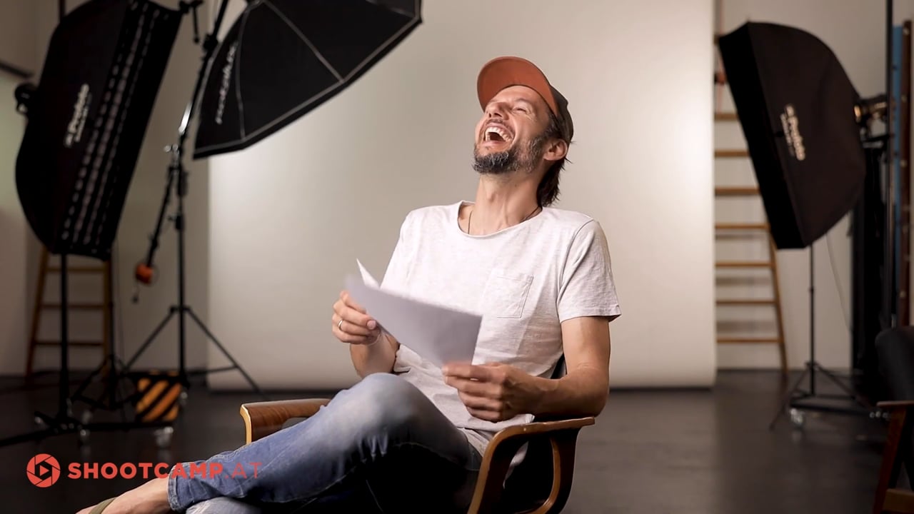 Mann im Studio lacht, hält Papier in Hand, trägt rote Mütze und weißes T-Shirt