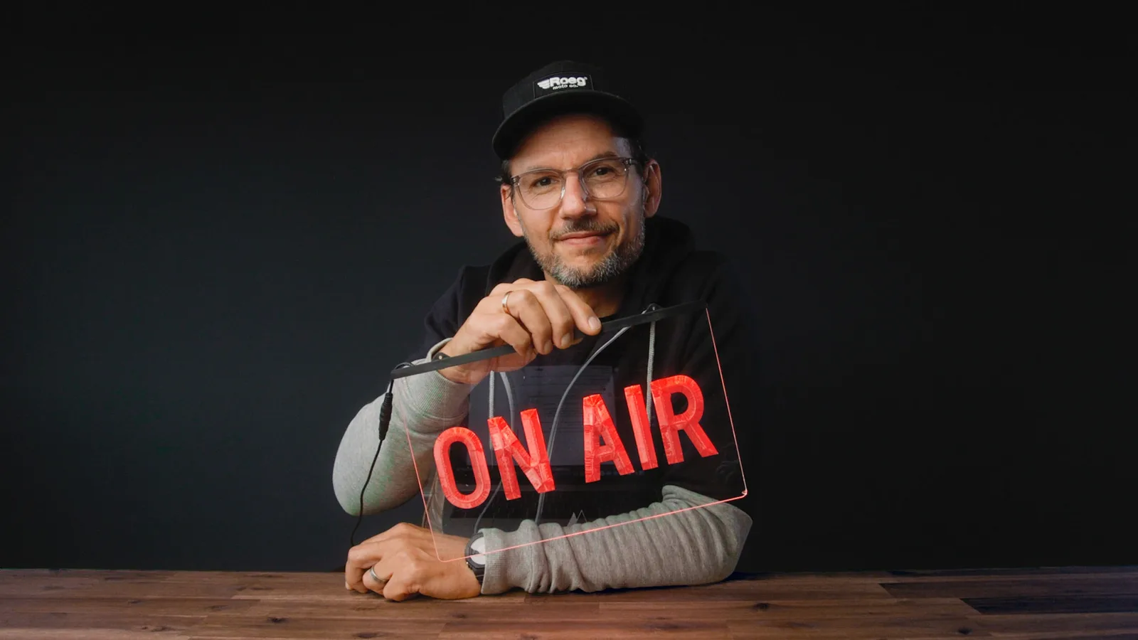 Mann mit Mütze und Brille hält Neon-Schild 'ON AIR' im Studio